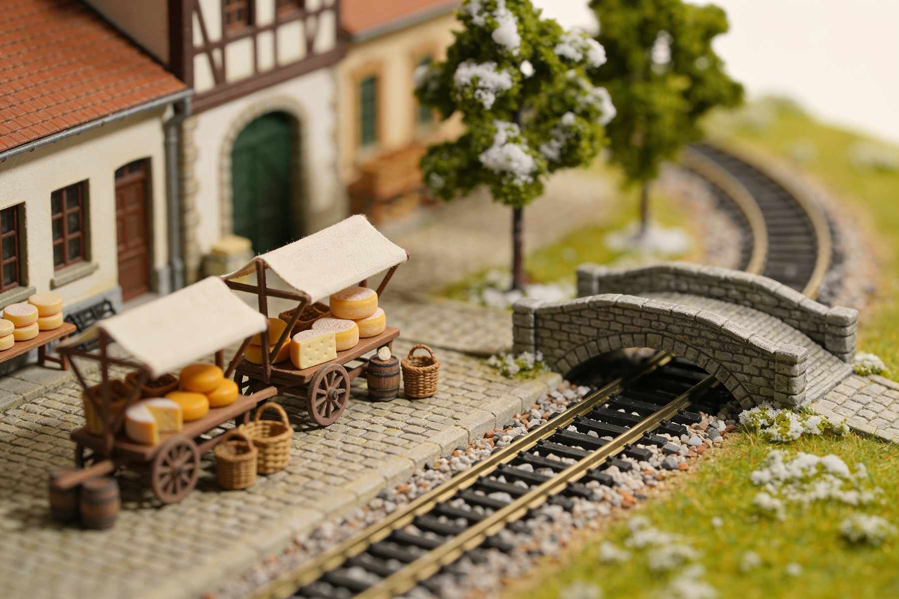 A tiny market square beside the railway with stalls of cheese wheels and baskets, cobbles and a stone bridge over the track